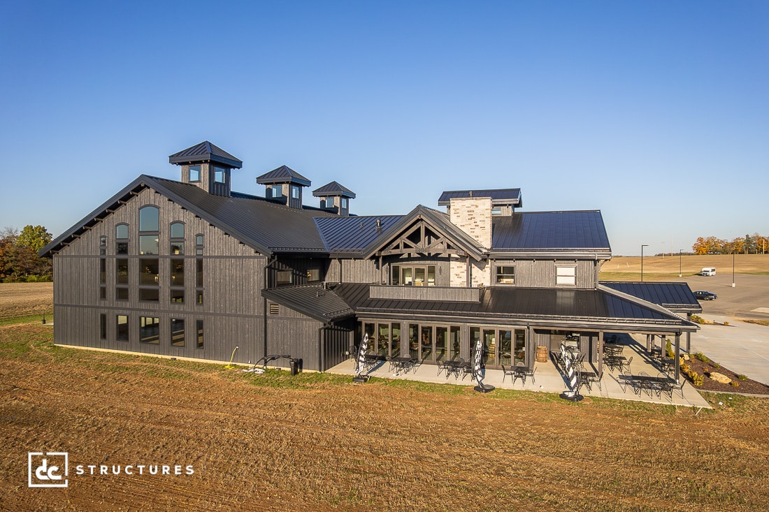 A large modern barn-style building with dark exterior, gabled roofs, and tall windows sits on a brown field under a blue sky.