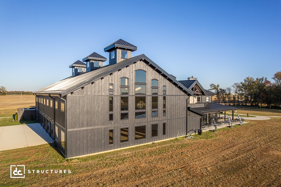 A large modern barn-style building with dark gray siding, tall windows, and multiple cupolas sits on a grassy lot by a paved driveway. Trees and open fields are in the background.