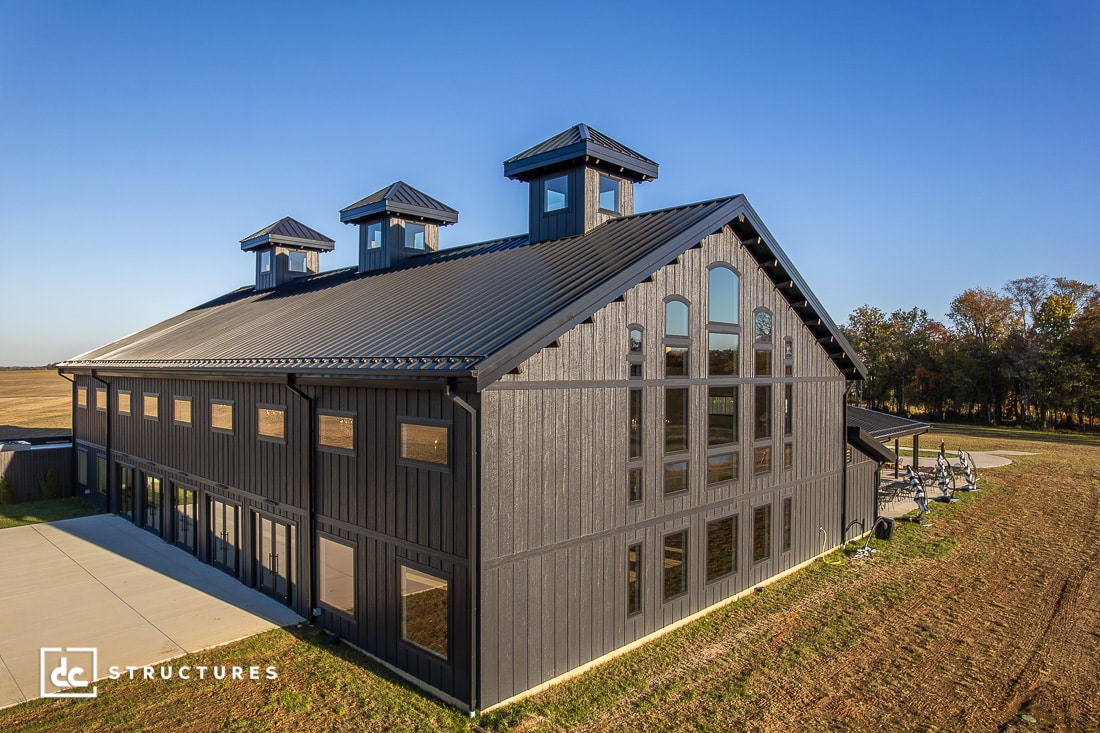 Large modern barn-style building with tall windows and three rooftop cupolas, set in a rural landscape with fields and trees. The structure has dark siding and a metal roof.