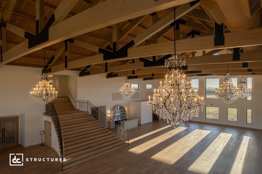 Large, elegant wooden hall with exposed beams, grand staircase, and three crystal chandeliers. Sunlight streams through tall windows.