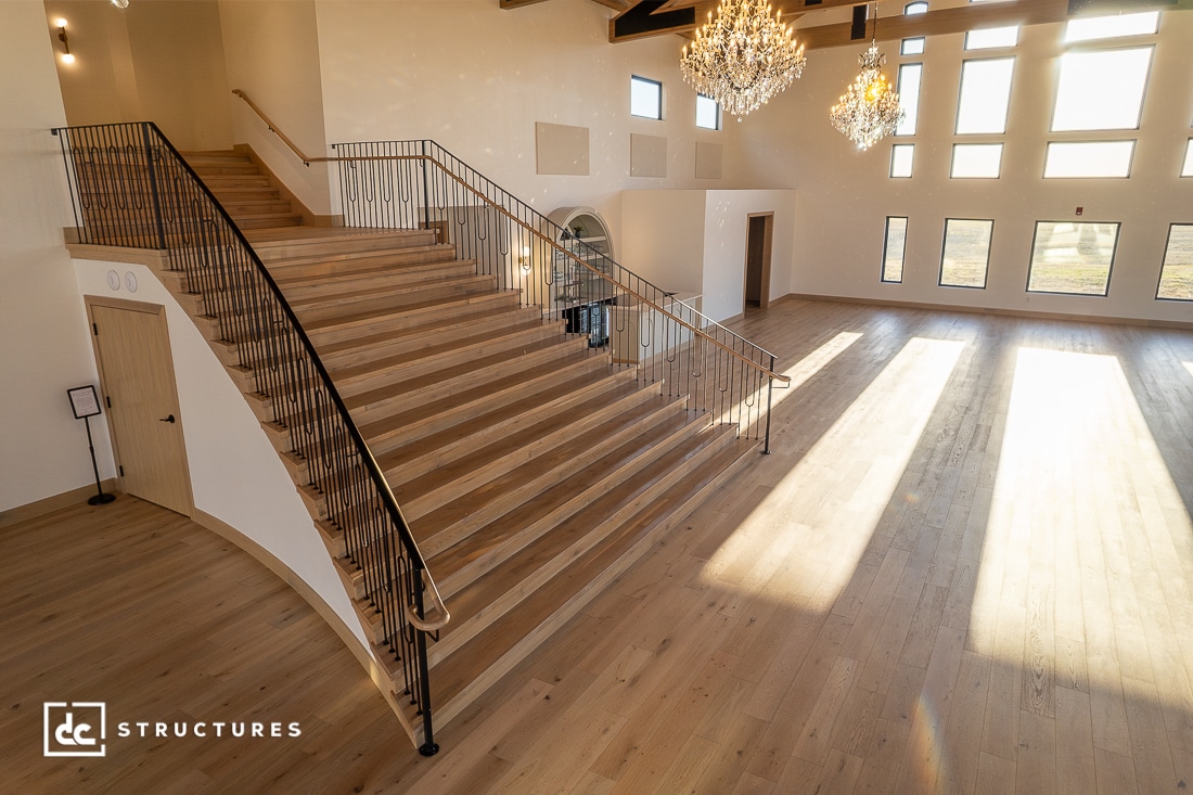A spacious, sunlit interior with large windows, a grand wooden staircase with black railings, elegant chandeliers, and light wood flooring.