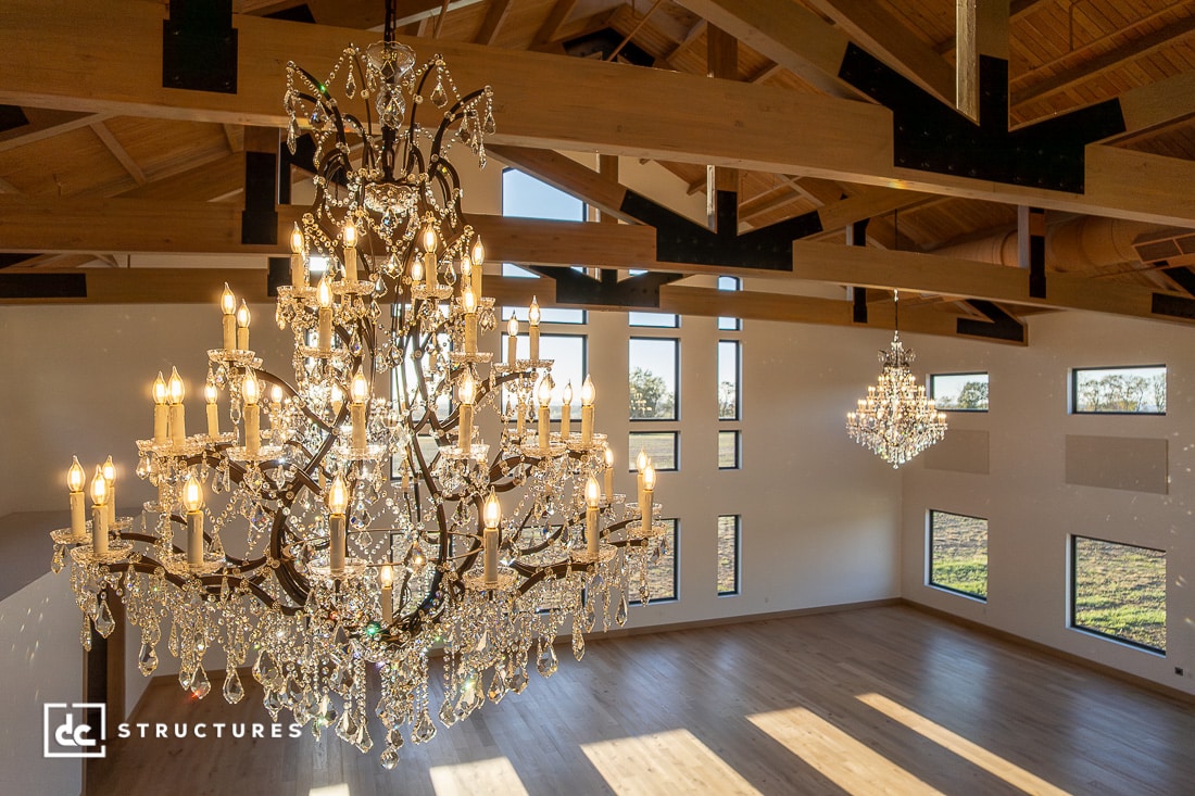 A large, ornate crystal chandelier hangs from a high wooden ceiling in a sunlit room with tall windows and exposed beams.