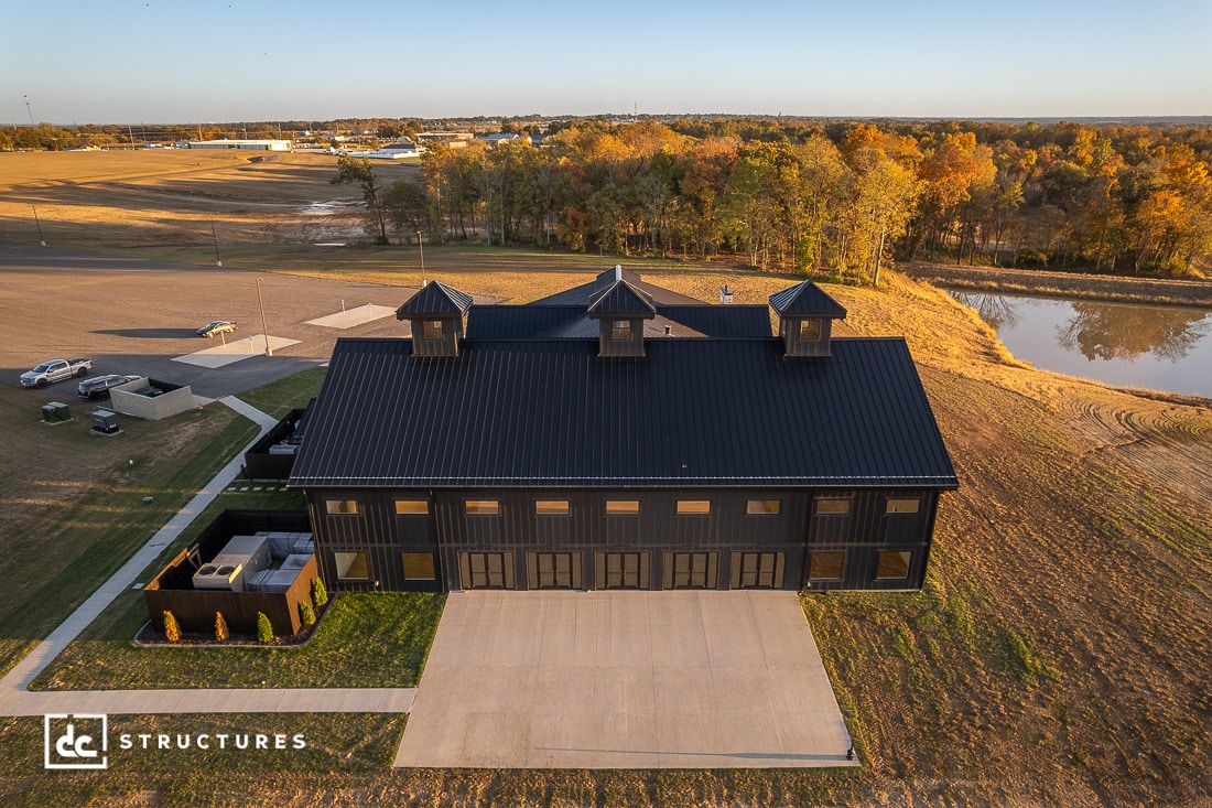 A large, modern black barn with three cupolas sits on a grassy lot by a pond, surrounded by autumn trees and a parking area.