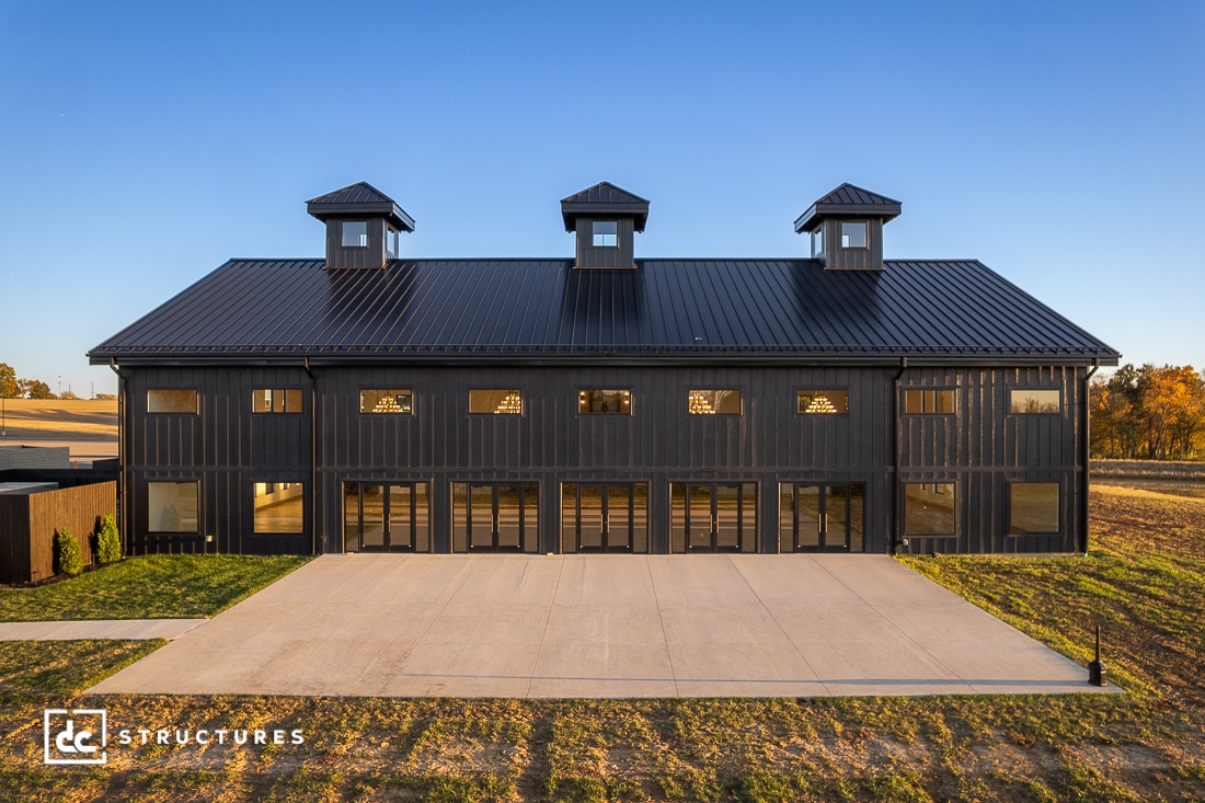 A large modern black barn with three cupolas, tall windows, and glass doors sits on a concrete pad amid open land under blue sky.
