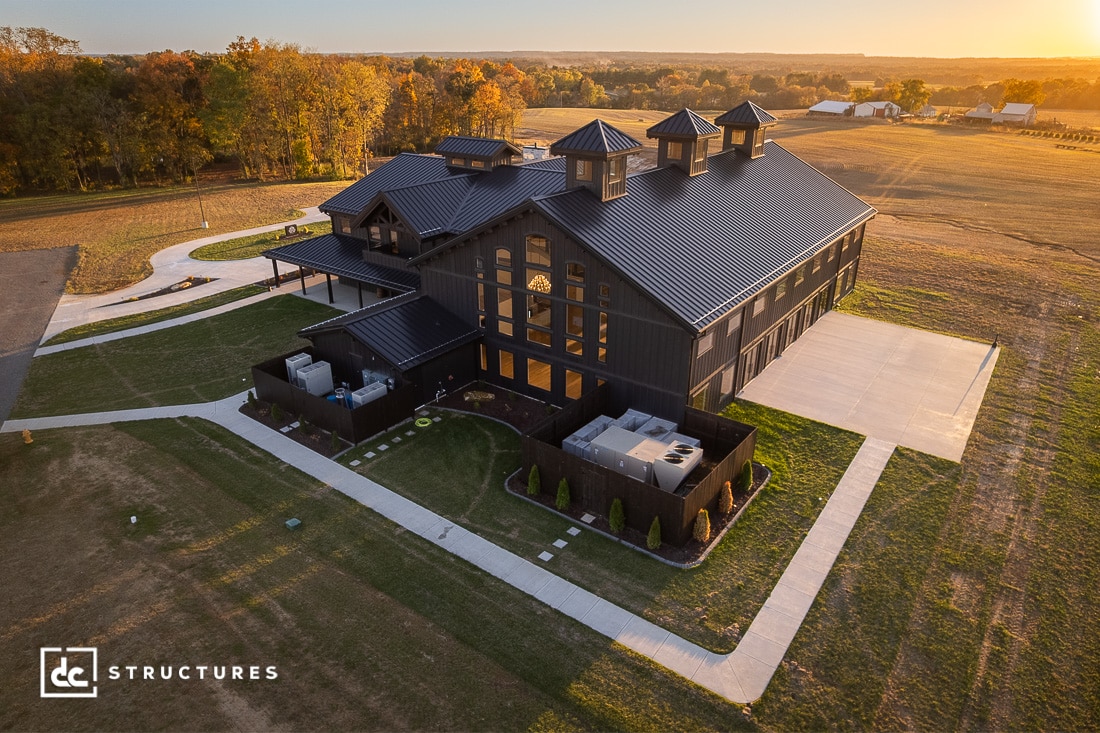 Aerial view of a large, modern black barn-style building with multiple cupolas, surrounded by grass, trees, and concrete walkways at sunset.