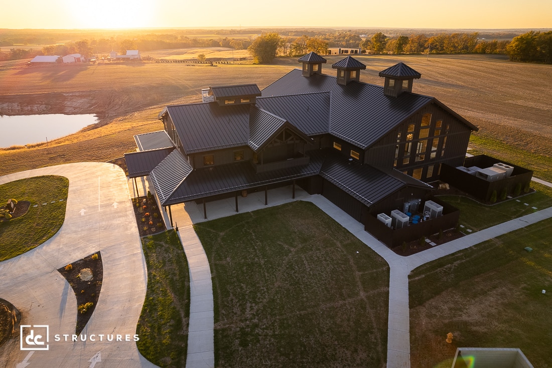Aerial view of a large modern barn-style house with black metal roofing, surrounded by open fields and a pond at sunset.