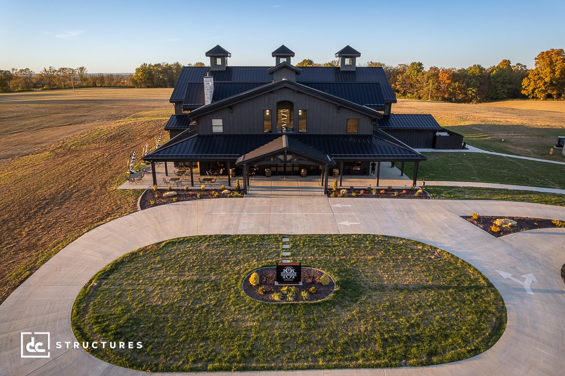 A large, modern black barn-style building with multiple cupolas sits amid open fields. A circular driveway and manicured landscaping frame the entrance. Trees line the horizon under a clear sky.
