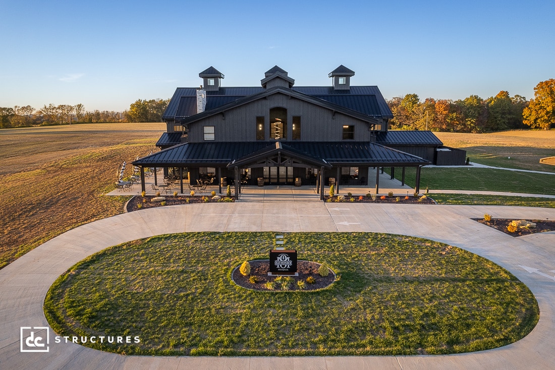 A large modern black barn-style building with a wraparound porch, spacious green lawns, trees behind, and a circular driveway in front.