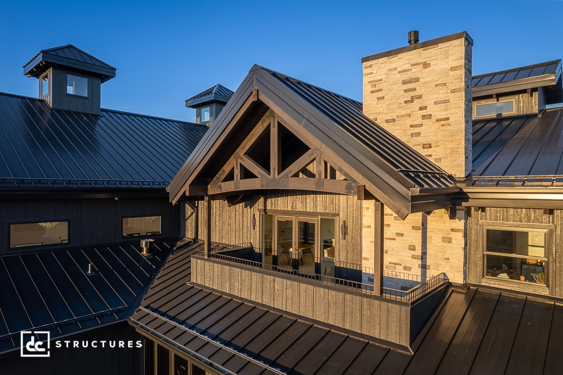 A modern house with dark metal roofing, a stone chimney, and a covered balcony. Exposed beams at sunset with a clear blue sky.