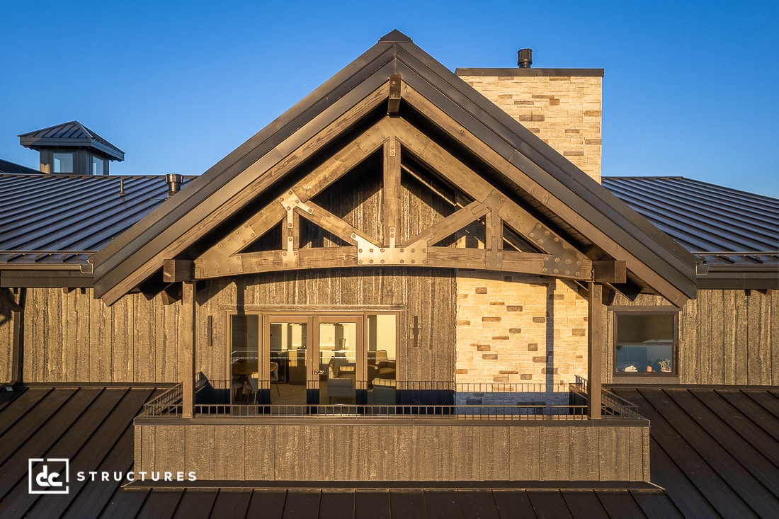 A close-up view of a modern house with a metal roof, exposed wooden beams, and a balcony. Warm light shines from the interior windows, and a stone chimney is visible in the background.