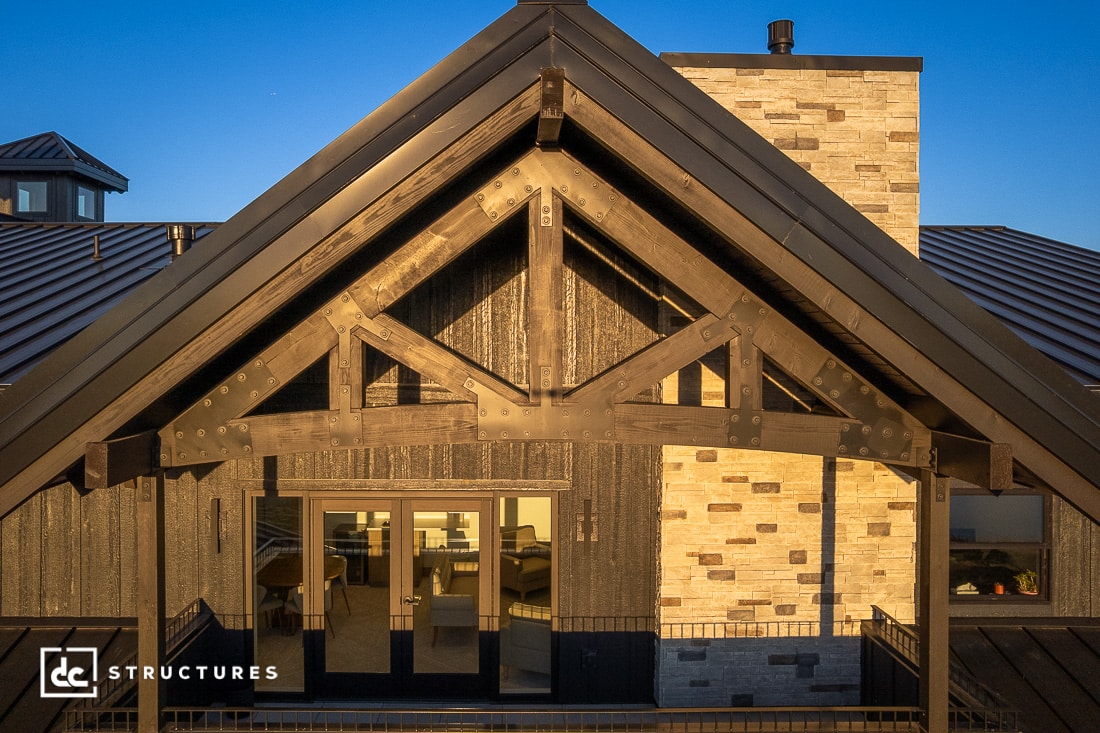 Exterior view of a modern house with a black metal roof, exposed timber trusses, large glass doors, and beige stone chimney at sunset.