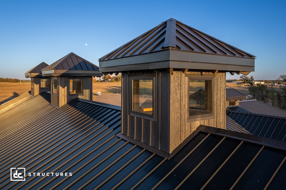 View of a modern metal roof with two cupolas featuring large windows in a rural setting at sunset, with fields and distant trees.
