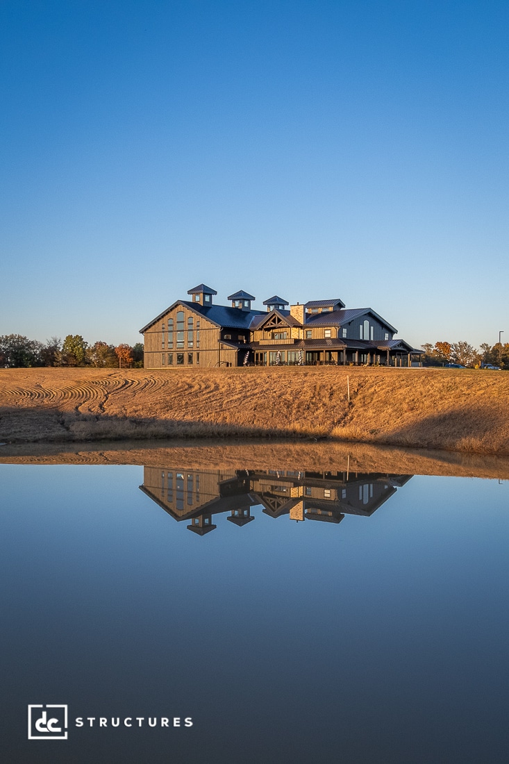 A large, rustic barn-style building sits atop a grassy hill, reflected in a calm pond under a clear blue sky.