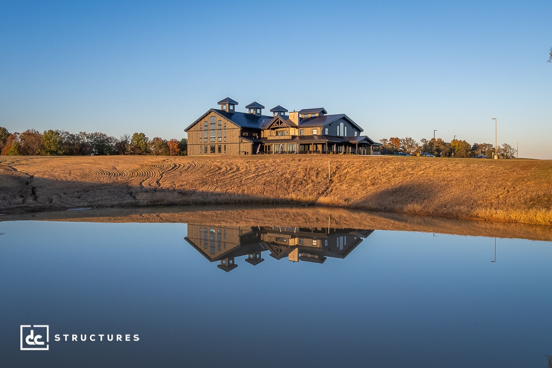 A large modern barn-style building sits on a grassy hill at sunset, reflected clearly in a calm pond below; trees and a blue sky are in the background.