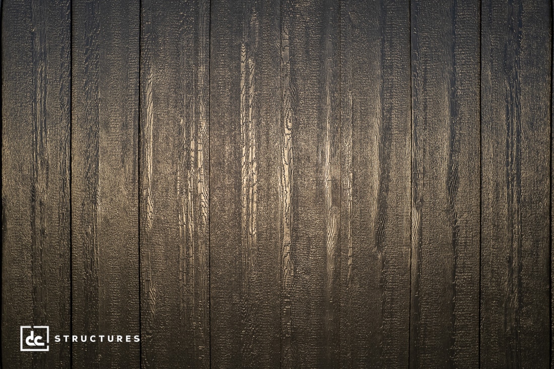 A close-up of vertically aligned wooden panels with a dark, textured finish. Light reflects off the surface, highlighting the wood grain.