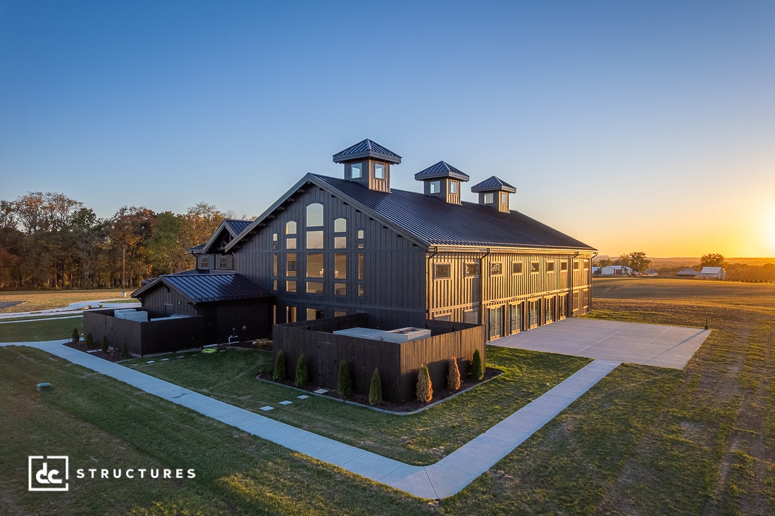 A large modern barn with dark wood siding, tall windows, and three cupolas sits on manicured grass with paved driveways at sunset.