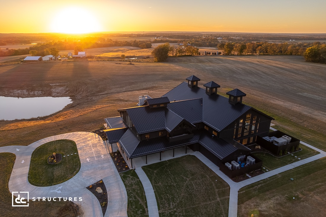 A large modern barn-style house with a dark roof sits on farmland at sunset, surrounded by fields, a pond, and distant outbuildings.