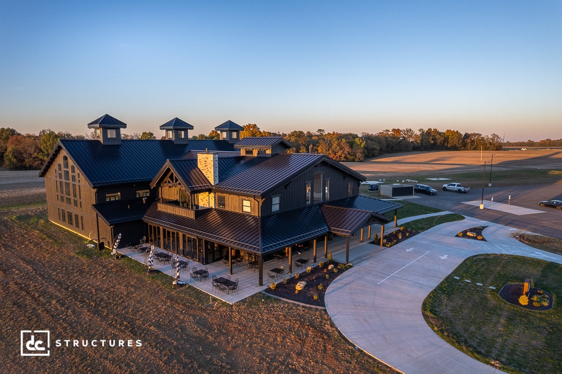 A large modern barn-style building with black metal roofing sits in a rural landscape at sunset, with gabled roofs and outdoor seating.