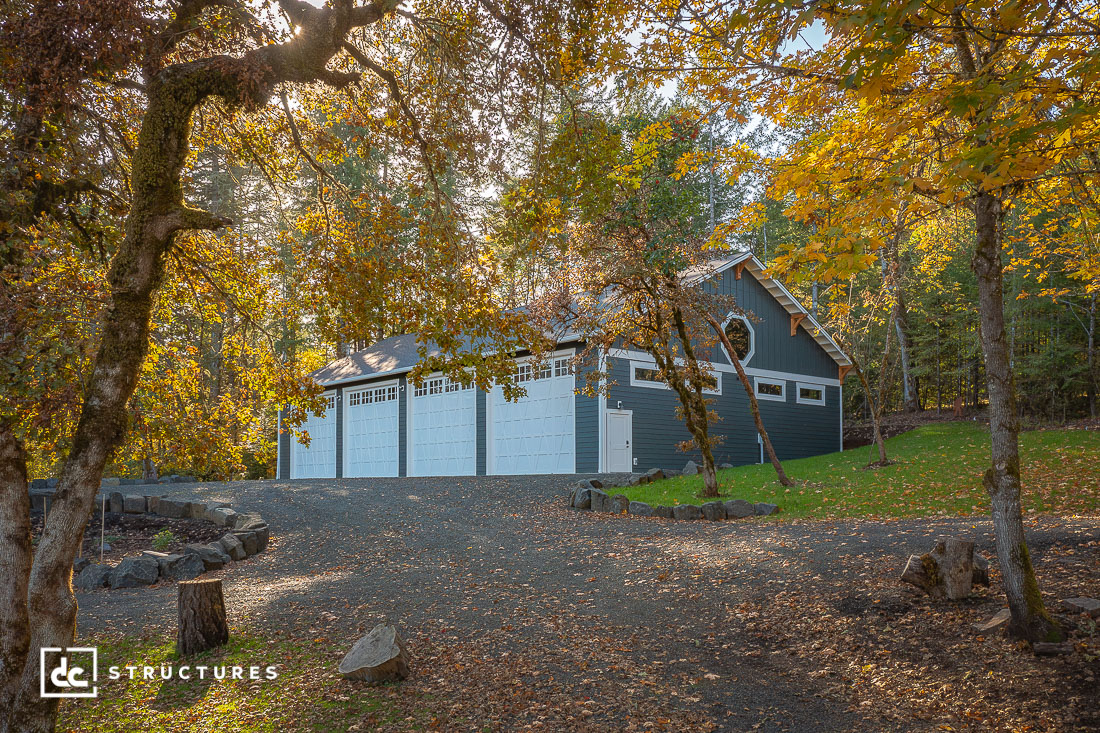 A large, dark gray garage with white doors sits among autumn trees with golden leaves. A gravel driveway leads up to the building.