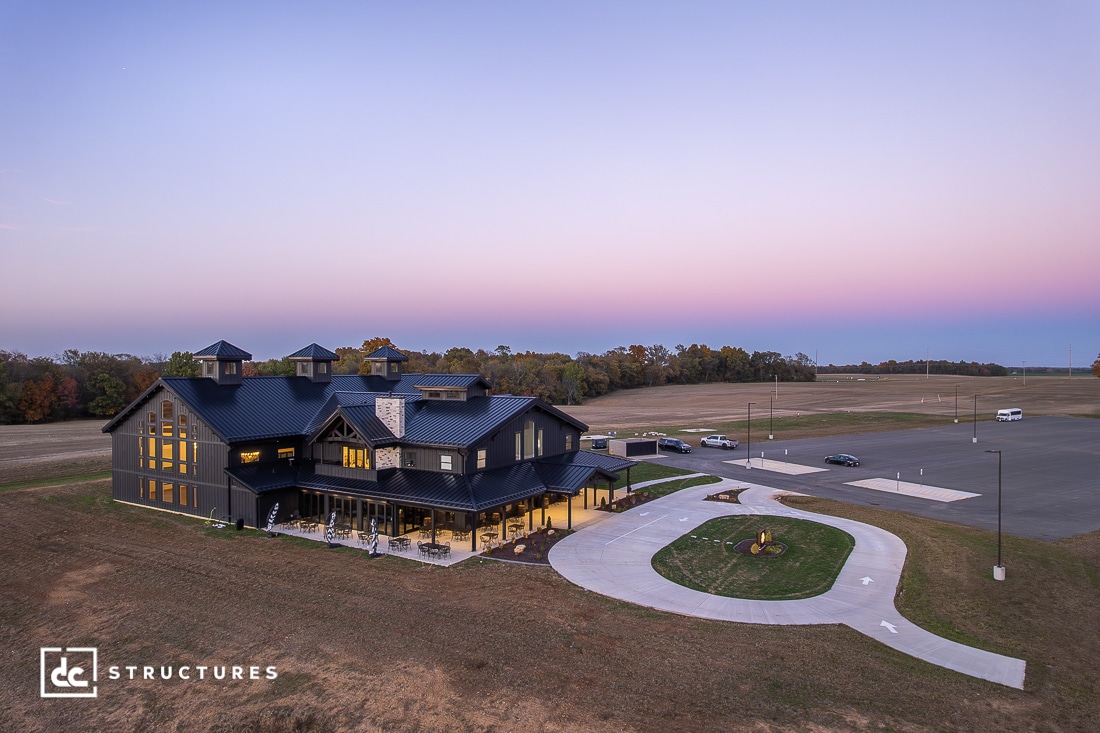 A large modern barn-style building with black exterior sits on open land at sunset, with a pink and purple sky overhead.