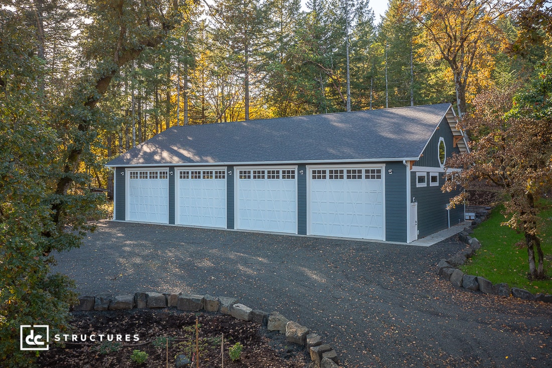 A large gray garage with four white doors stands surrounded by trees with green and autumn leaves. Gravel paves the driveway.