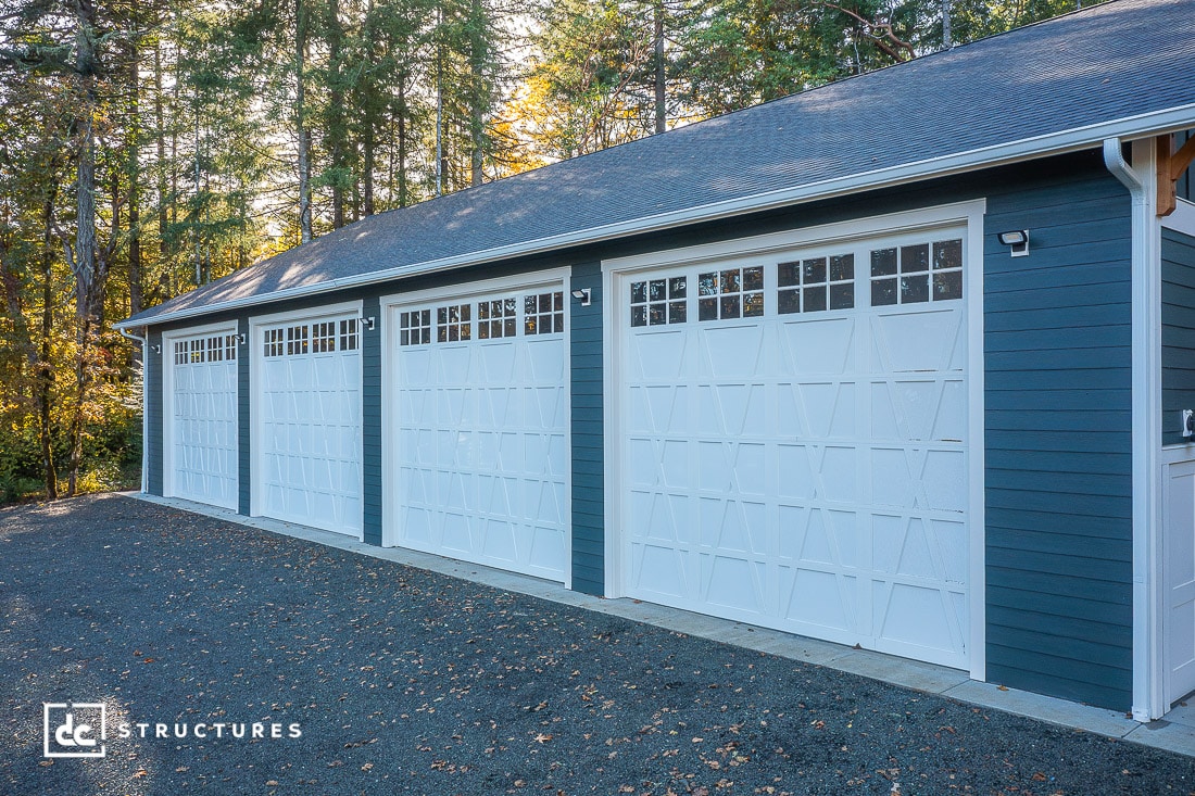 A row of four large white garage doors with decorative windows on a blue building, surrounded by tall trees and sunlight filtering through the forest. The ground is covered with gravel.