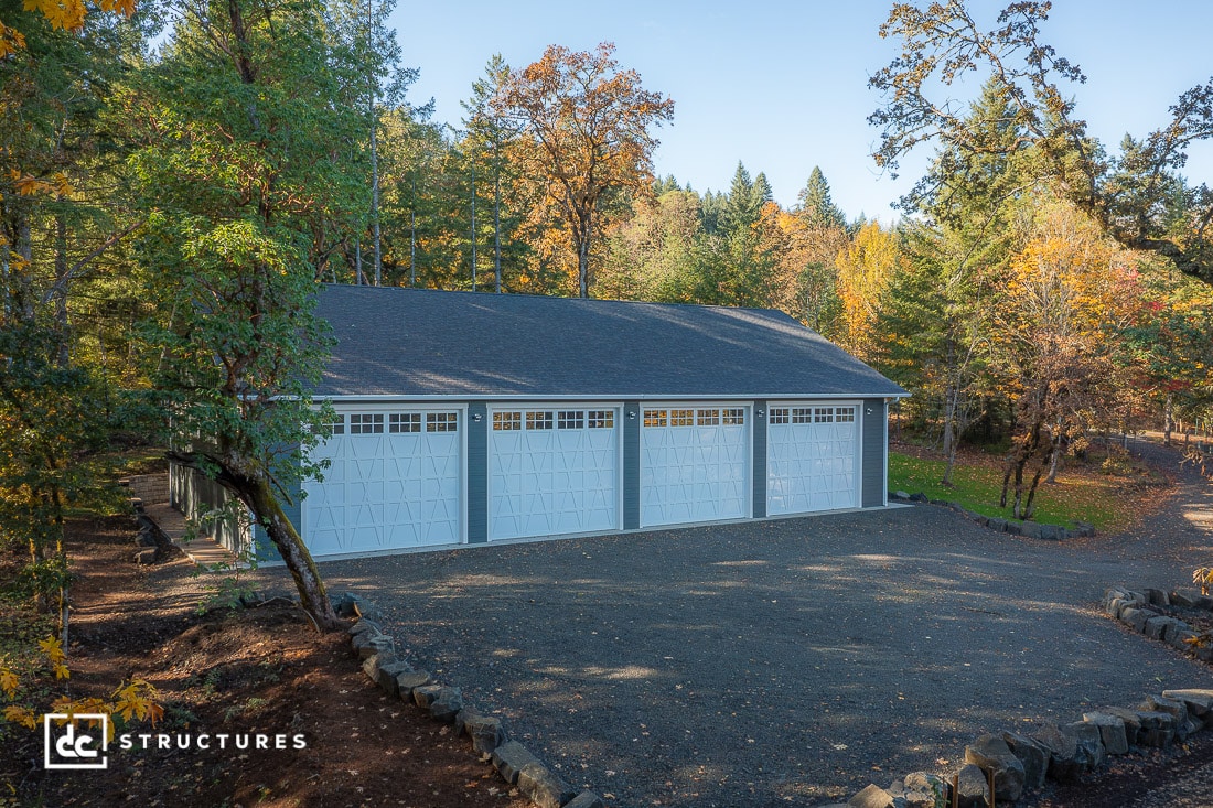 A large white four-car garage with light blue doors is surrounded by autumn trees under a clear sky, with gravel in front.