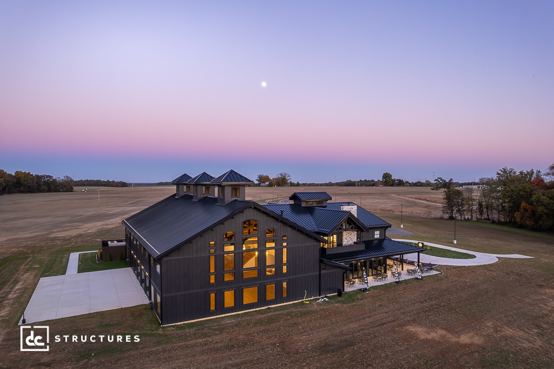 A modern barn-style building with large windows and black siding stands on a wide, open field at sunset. The sky is purple and pink, with a visible moon.
