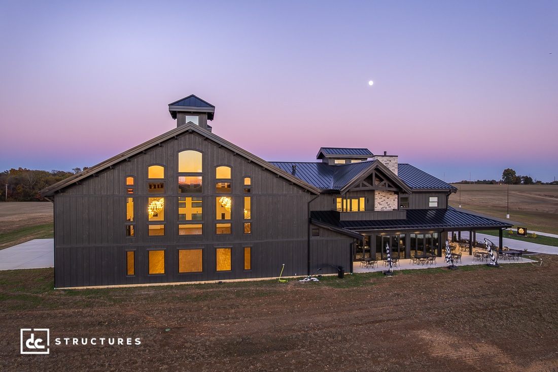 A modern barn-style building with large windows and outdoor seating stands on open land at dusk under a clear sky with a visible moon. Lights glow warmly inside.