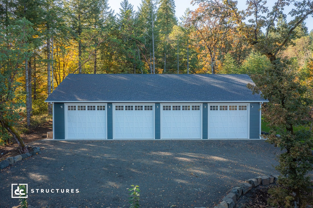 A large blue four-car garage with white doors on a gravel driveway, surrounded by autumn trees and a dark shingle roof.