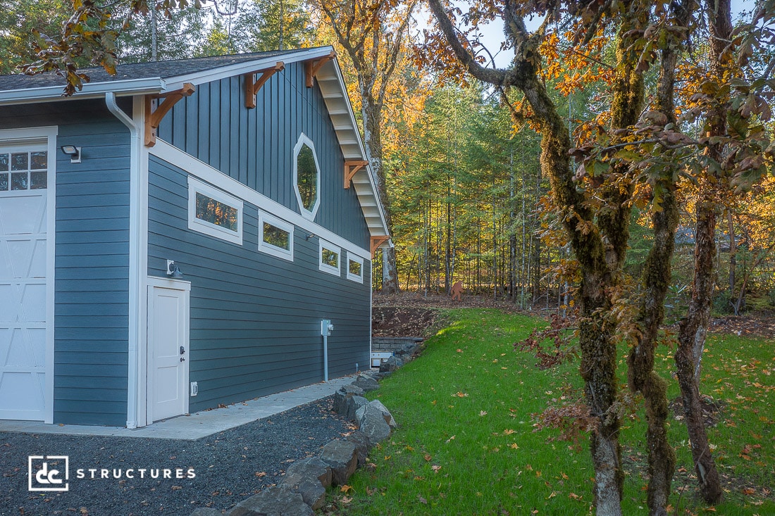 A modern blue barn with white trim sits by a grassy yard and autumn trees, surrounded by forest. A gravel path runs beside it.