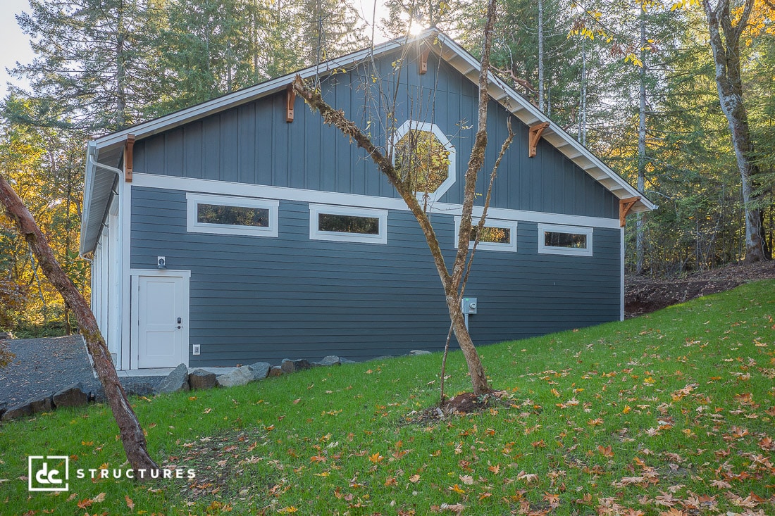 A blue and gray barn-style building with white trim, rectangular windows, and a side door sits on a grassy lawn among trees.