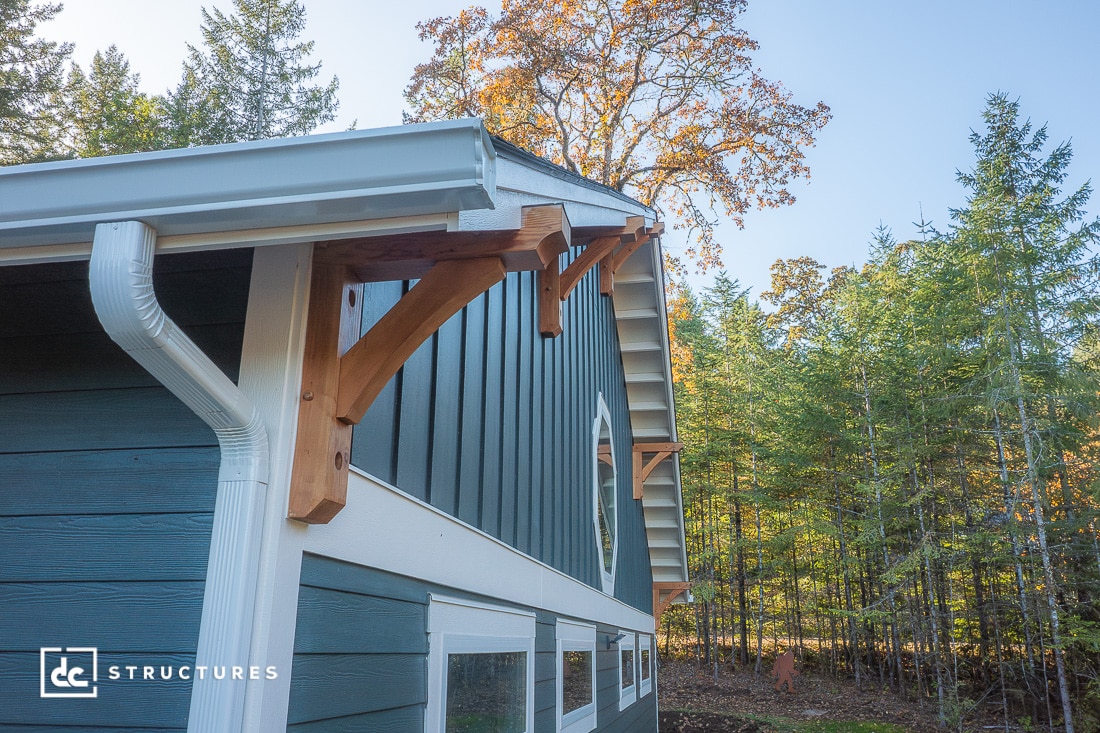 A close-up of a modern barn’s corner shows blue siding, white trim, decorative wooden brackets, and a metal roof with trees and sunlight in the background.
