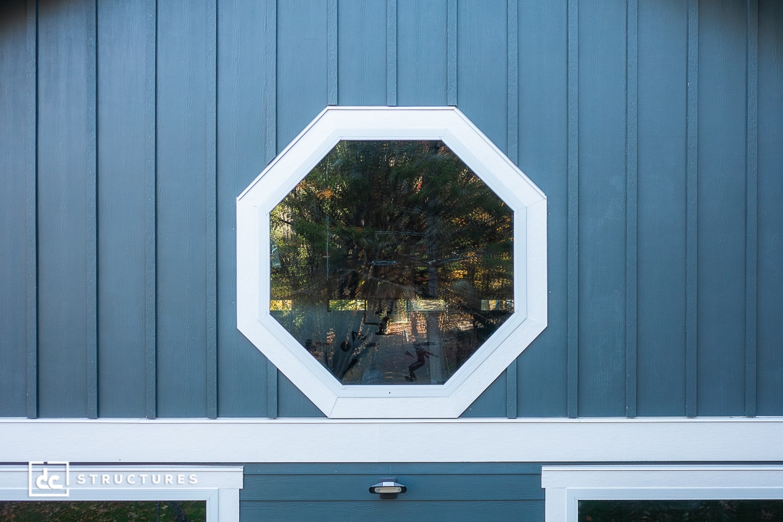 An octagonal window with a white frame is set into a blue vertical-paneled wall. The window reflects the image of a tree.