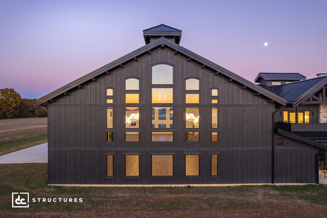A large modern barn-style building with tall, symmetrical windows is illuminated at dusk. A purple sky and moon are visible.
