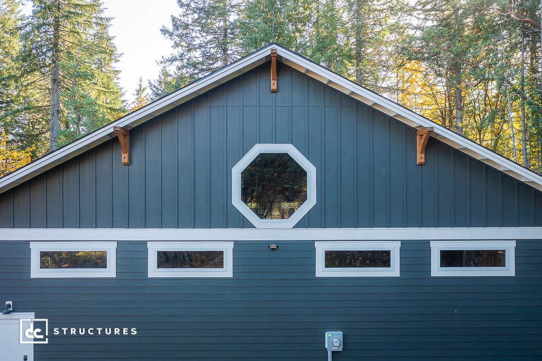 A blue-gray building with white trim, an octagonal window near the peak, and four windows below, set against tall evergreens.