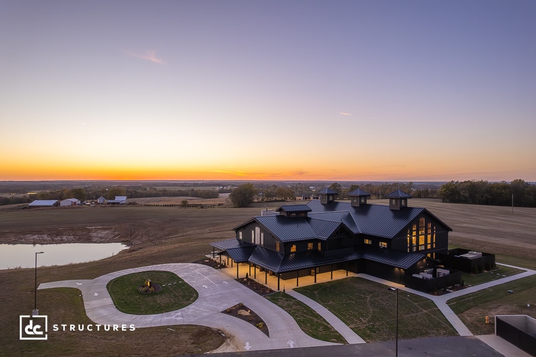 A modern black barn-style building with large windows sits amid open fields at sunset, with a circular driveway and small pond.