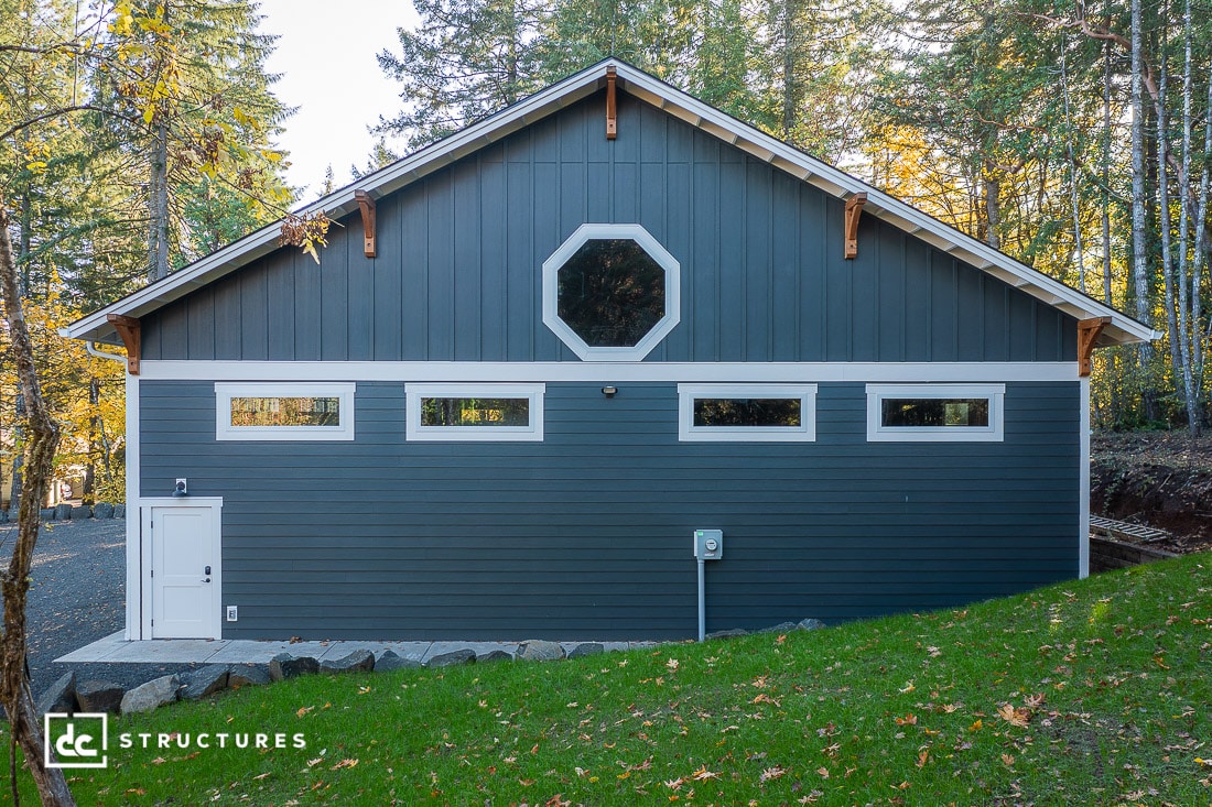 A blue and gray barn-style building with four narrow rectangular windows and an octagonal window above, surrounded by trees.