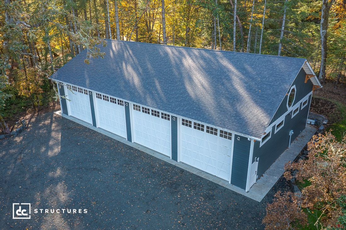 A large, dark blue garage with four white doors and decorative windows sits surrounded by trees on a gravel lot. The building features a peaked roof.
