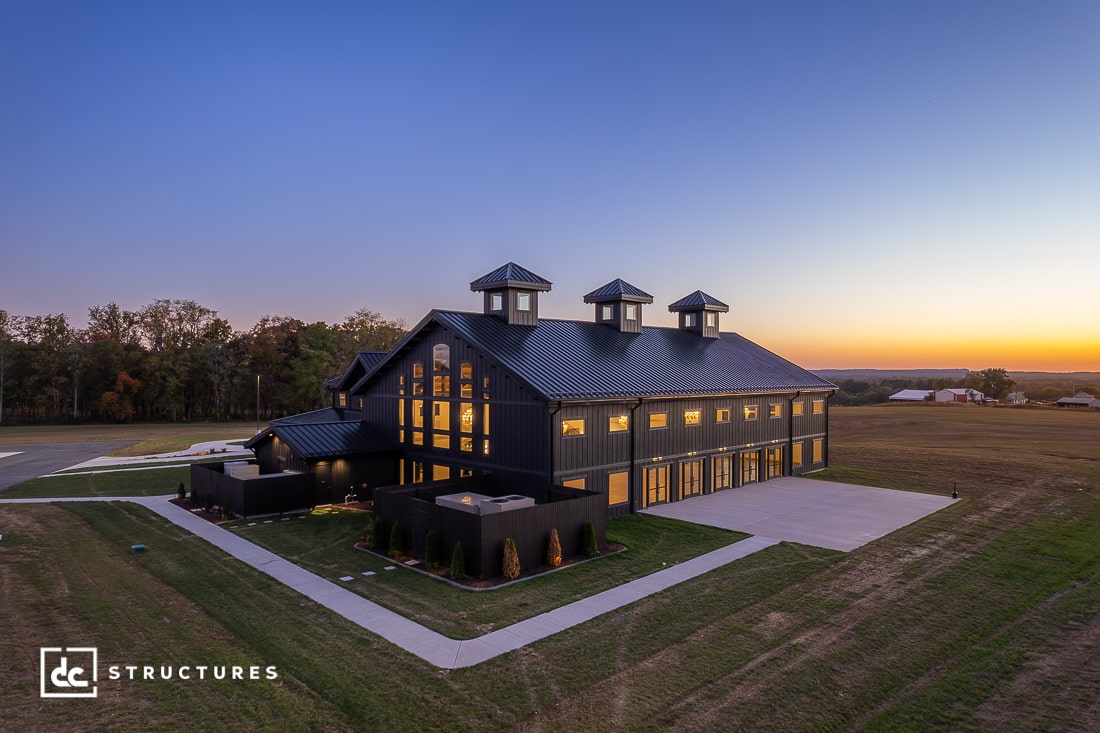 A large, modern black barn-style building with numerous windows and cupolas is illuminated at sunset, surrounded by grass, trees, and a concrete driveway.