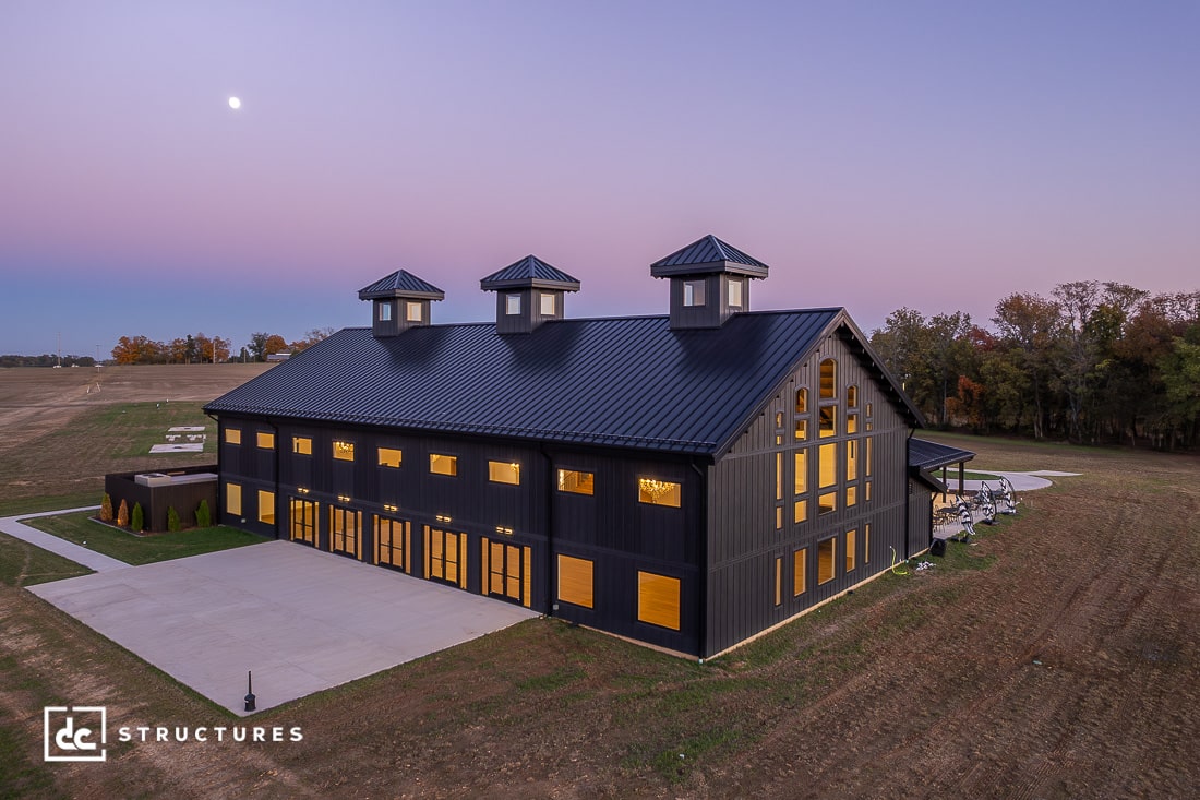 A large black barn with three cupolas and many lit windows stands on a concrete lot at dusk under a clear sky with a visible moon.