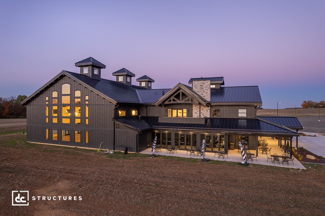 A large modern barn-style building with dark siding, tall windows, and cupolas sits on an open lot at dusk with warm lights glowing.