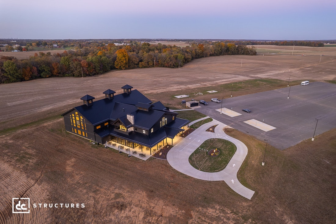 Aerial view of a modern barn-style building with black roof, fields and trees. Curved driveway, large empty parking lot on the right.