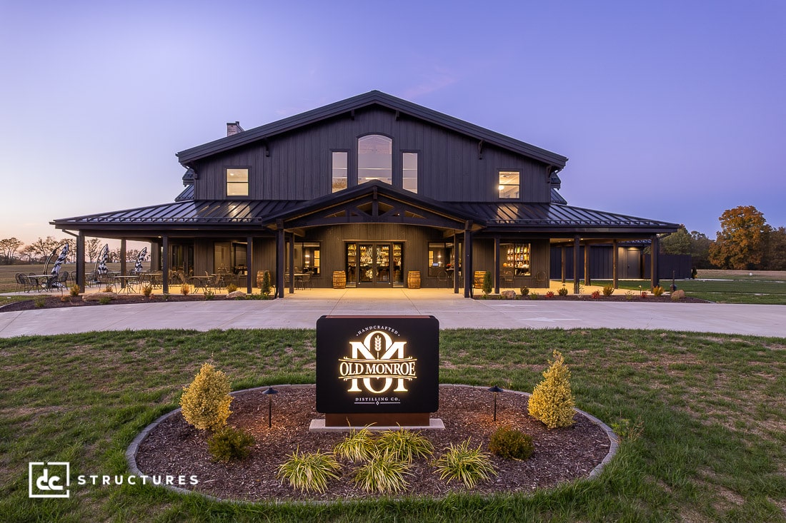 A large, modern barn-style building with black siding and expansive windows is lit up at dusk. Manicured landscaping lines the entrance walkway.