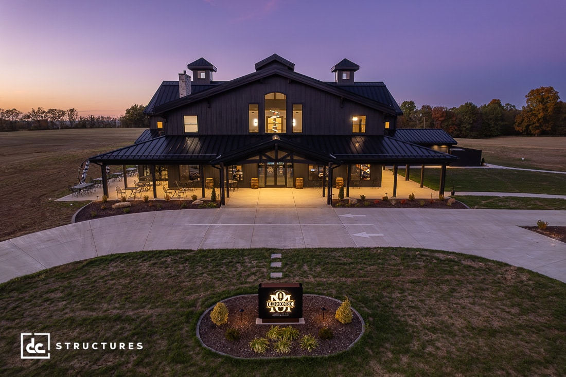 A large, modern barn-style building with lit windows and cupolas stands in open fields at dusk, with a curved driveway in front.