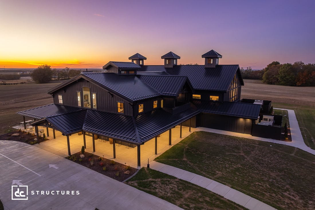 A large, modern black barn with multiple cupolas is illuminated at sunset, surrounded by open fields and a driveway.