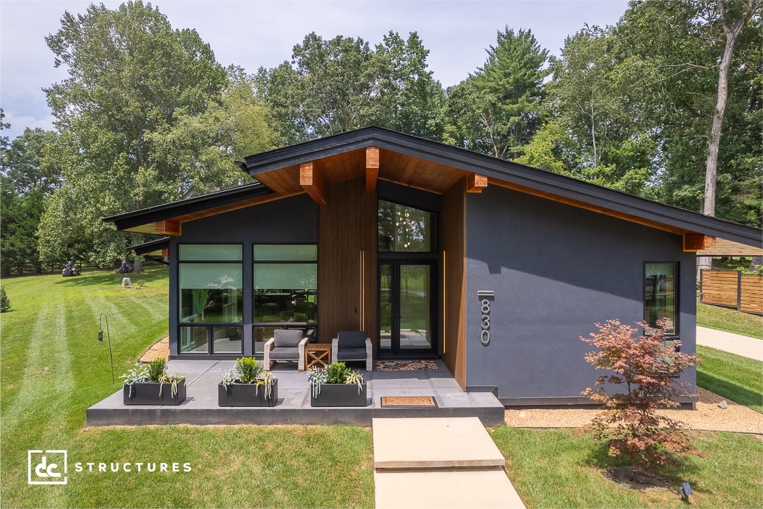 A modern single-story house with dark exterior walls, large windows, a covered porch, sloped roof, green grass, trees, minimal landscaping.