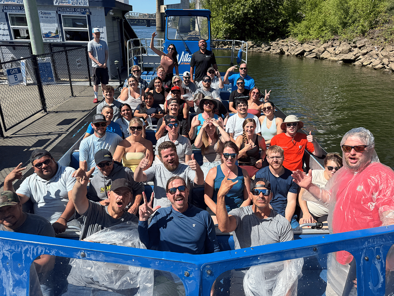 A group of smiling people sit on a blue boat, posing for a photo with peace signs and thumbs up by a calm river on a sunny day.