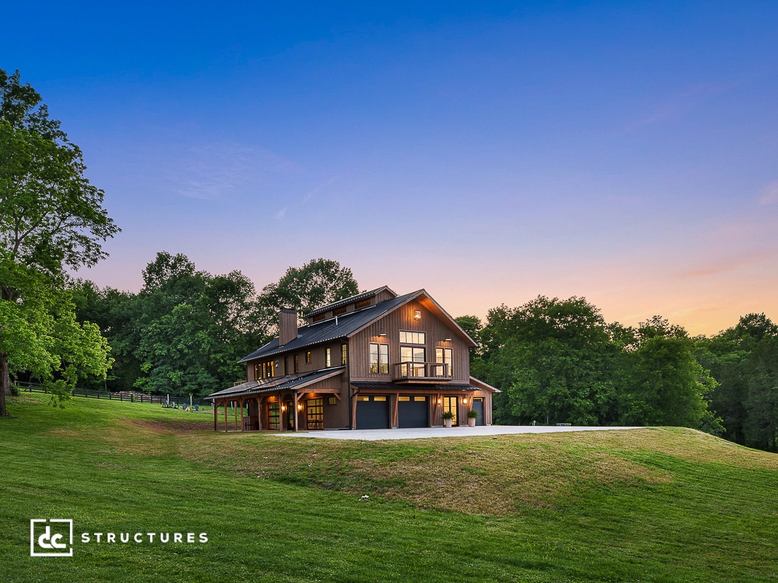 A modern barn-style home with large windows and exterior lights sits on a grassy hill, surrounded by trees at sunset.