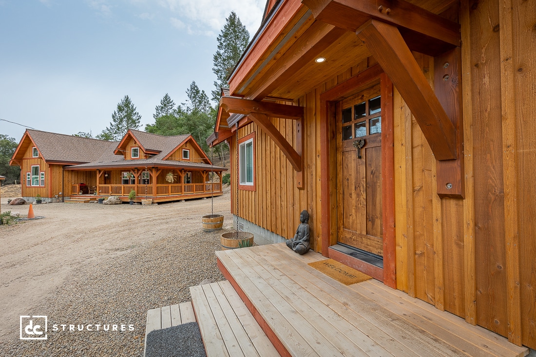 A wooden house with a covered porch and a Buddha statue by the front door sits in the foreground; another similar wooden building stands in the background among trees and gravel.
